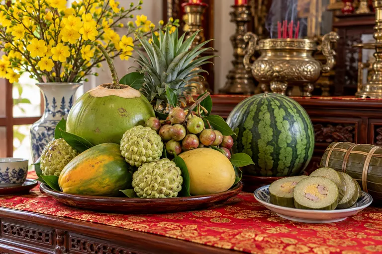 Mâm ngũ quả miền Nam: trái dừa, mãng cầu, đu đủ, xoài và chùm sung, phía sau là trái thơm (dứa); bên cạnh có trái dưa hấu, đĩa bánh tét cắt khoanh và bình hoa mai vàng.